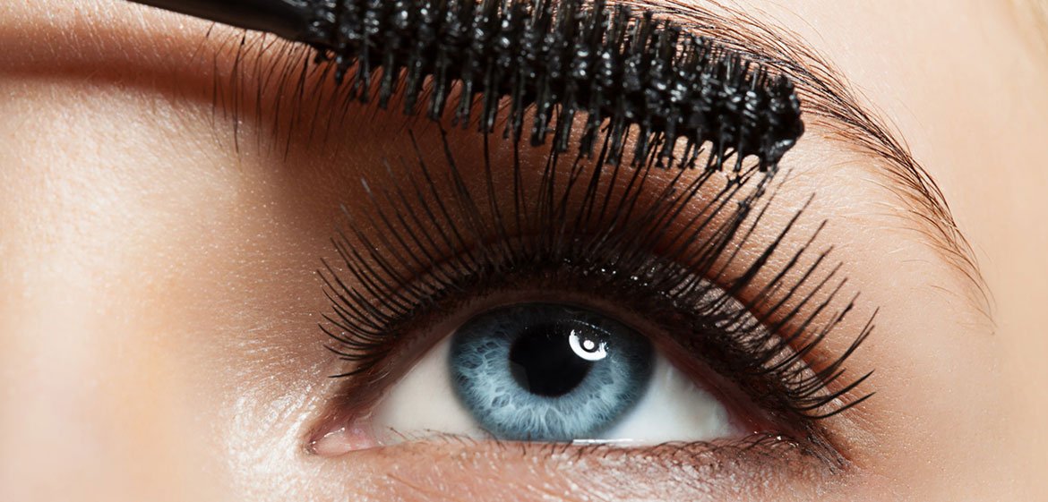 Nahaufnahme eines blauen Frauenauges mit langen, schwarzen Wimpern, die von einer Mascara-Bürste getuscht werden.